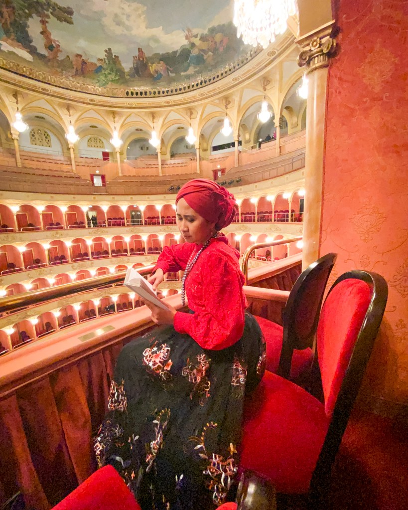A woman dressed in traditional attire with a red top and a patterned skirt sits reading a book in a vintage theatre. The ornate ceiling and plush red seating create an elegant ambiance.