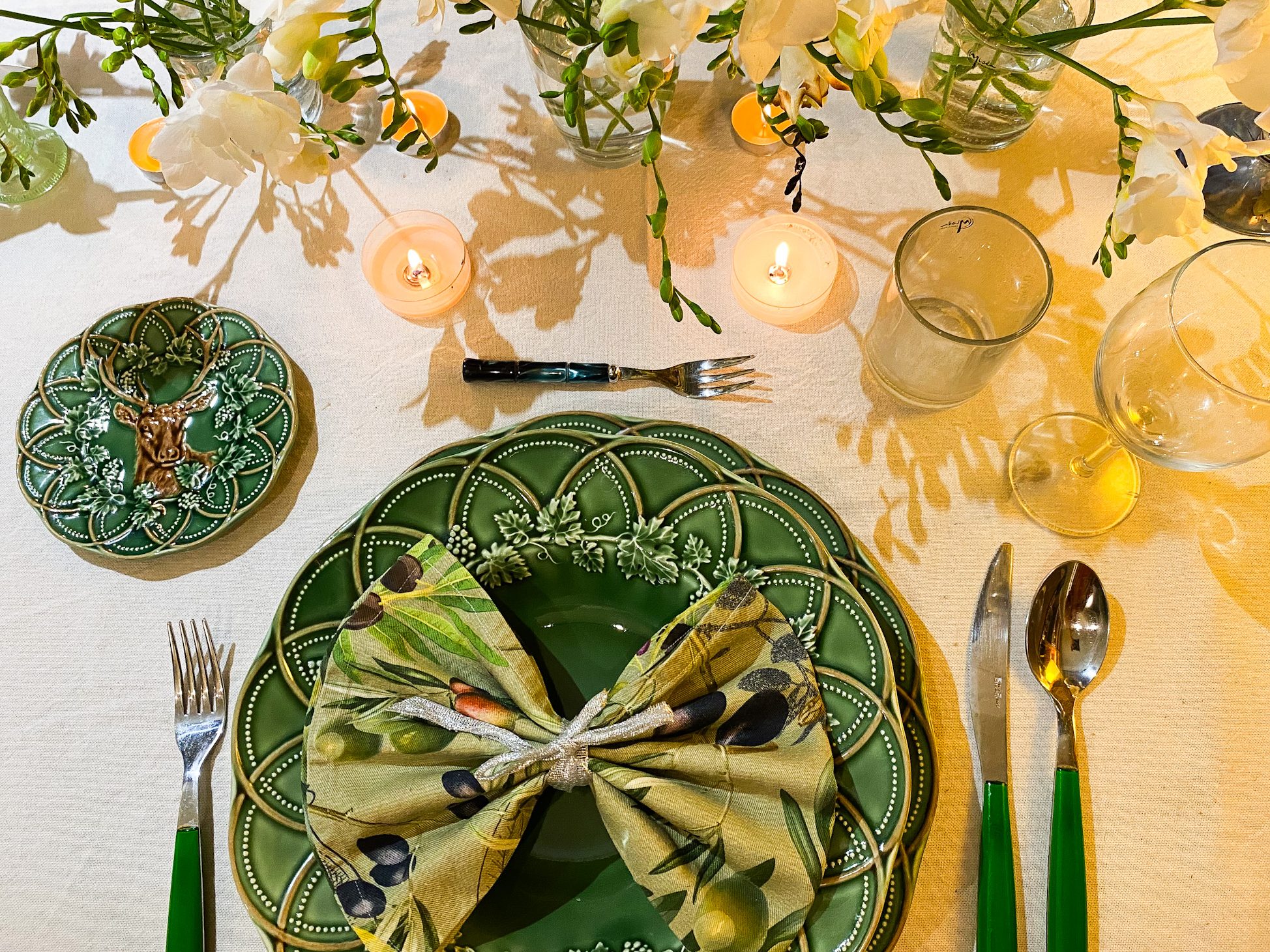 A beautifully set dining table featuring a green decorative plate with an olive-patterned napkin tied in a bow, a smaller decorative plate with a deer design, a fork, a knife, and a spoon with green handles. There are glassware, small candles, and floral arrangements in the background.