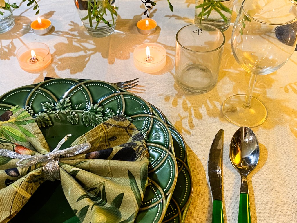 A beautifully set dinner table featuring a green plate with a decorative pattern, topped with an olive-themed napkin tied with a silver ribbon. The table is adorned with candles and decorative glasses amidst foliage in vases.
