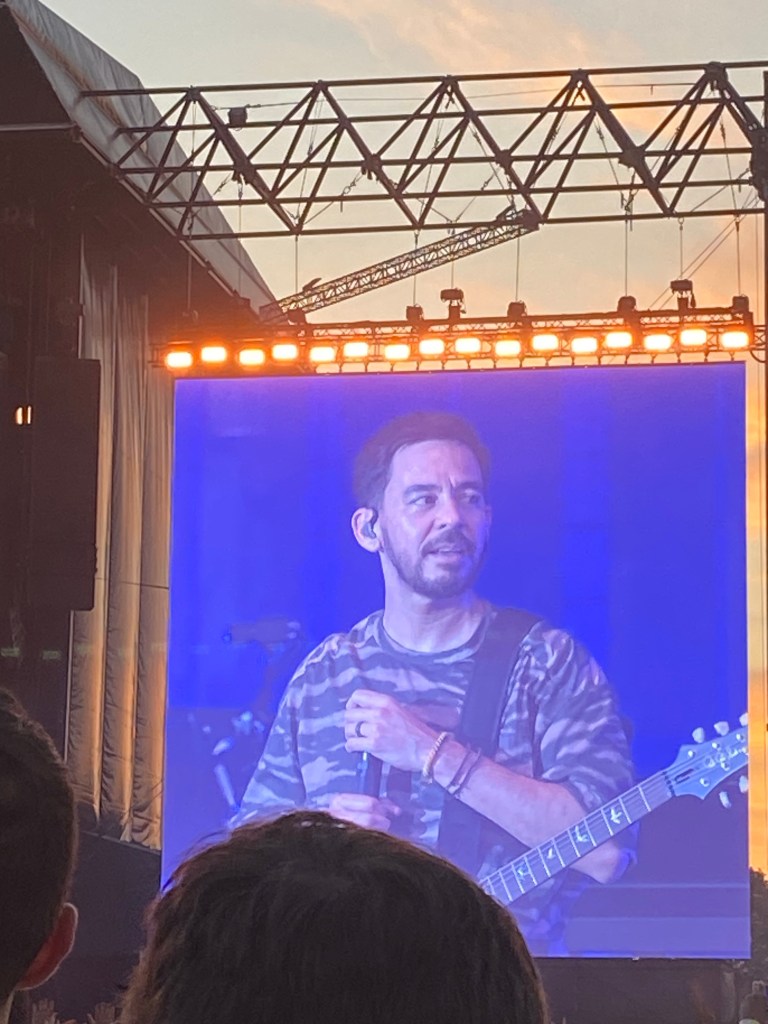 Mike Shinoda of Linkin Park on stage wearing a camouflage shirt, playing guitar and speaking to the audience, with a vibrant backdrop and lights visible.