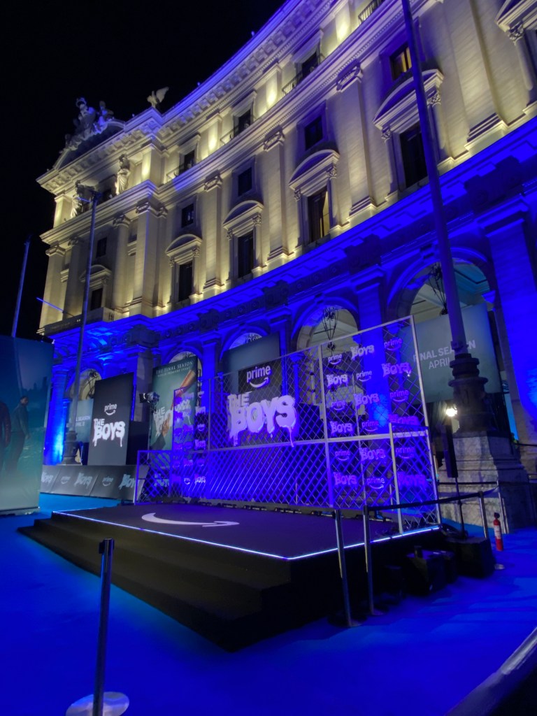 A night event setting featuring a stage with blue lighting and promotional banners for 'The Boys' in front of a historic building.