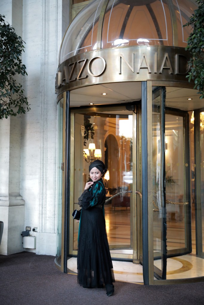 A woman wearing a black and teal dress poses elegantly near the entrance of a hotel with a glass revolving door, surrounded by decorative greenery.