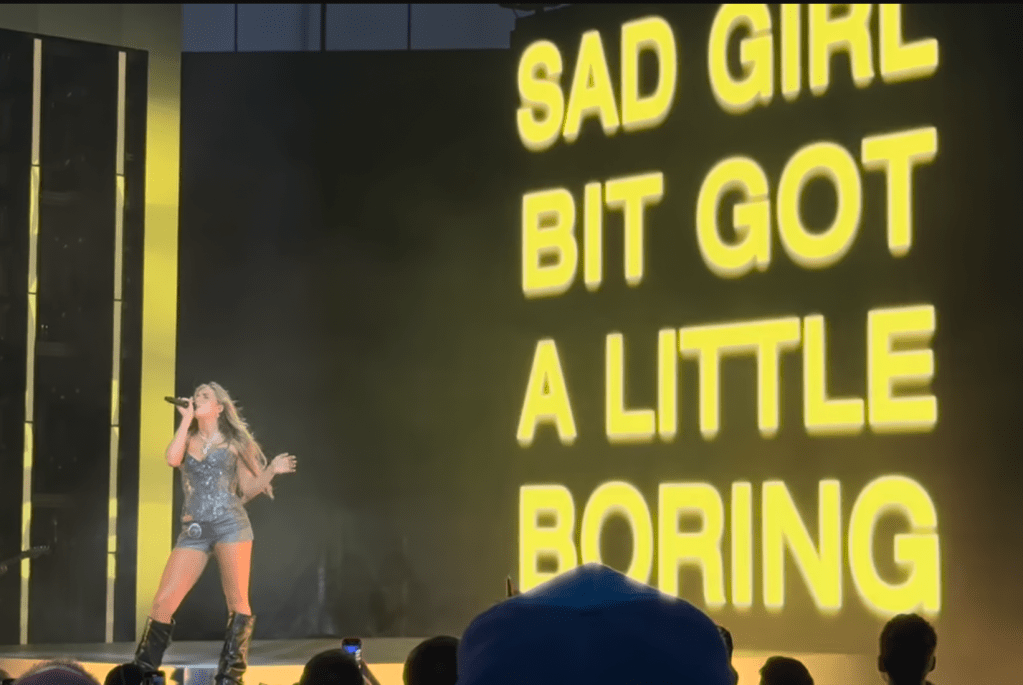 Tate McRae performing on stage with a microphone, wearing a sparkling outfit, in front of a large illuminated backdrop displaying the text 'SAD GIRL BIT GOT A LITTLE BORING'.