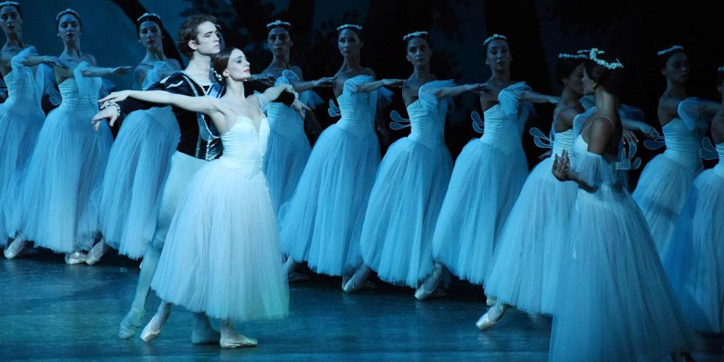 A group of ballet dancers performing on stage, wearing white tutus and pointe shoes, with one male dancer and a female dancer in the foreground executing a graceful pose.
