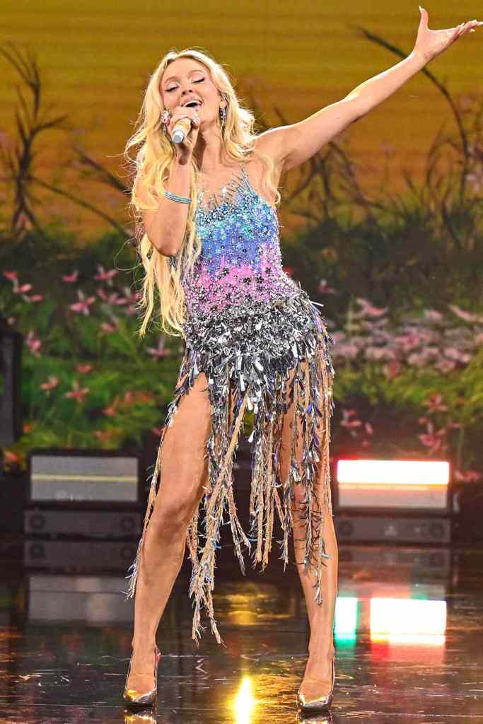 A singer performing on stage, wearing a sparkling multi-coloured dress with fringe details, and holding a microphone while gesturing expressively.