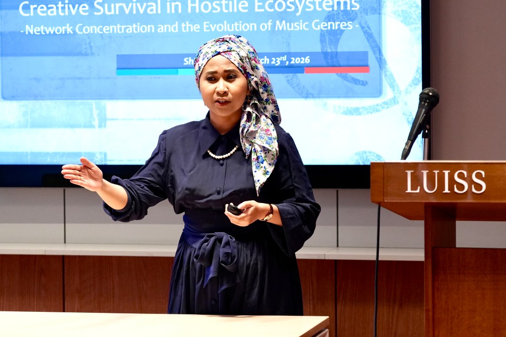 Shima Trisna speaking at the LUISS DIM Research Slam, gesturing as she presents research on the evolution of music genres, with a projected slide behind her.
